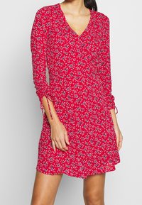 Rotes Blumenkleid mit langen Ärmeln, Wickeldesign und Raffung an den Bündchen. Aus leichtem, strukturiertem Stoff mit kleinen blauen Blumen.