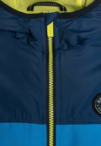 Granatowa kurtka z żółtym zamkiem błyskawicznym i czarnymi wykończeniami, z jasnoniebieską dolną częścią oraz okrągła naszywka z logo po prawej stronie.