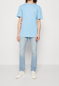 Camiseta de algodón azul claro con cuello redondo y mangas cortas, con un pequeño diseño gráfico blanco en el lado izquierdo del pecho, combinada con jeans claros y zapatillas blancas.