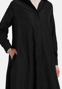 Manteau long noir en tissu lisse, avec patte de boutonnage avant, poches latérales et silhouette décontractée à manches longues.