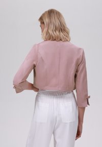 Veste en satin rose pâle, raccourcie, avec des manches trois-quarts et des accents en nœud. Associée à un pantalon blanc avec deux poches arrière.