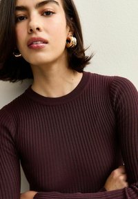 Maglione bordeaux a costine con scollatura a girocollo. Orecchini a cerchio dorati accentuano il look. La texture liscia conferisce un aspetto aderente.