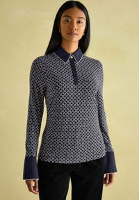 Marineblaues Langarmhemd mit weißem geometrischem Muster, Knopfdurchzugkragen und ausgestellten Manschetten. Glattes Gewebe mit kontrastierenden strukturierten Akzenten.