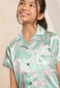 Camisa de pijama de satén verde menta con patrones de cinta blanca, manga corta, un bolsillo en el pecho y botones negros en la parte delantera.