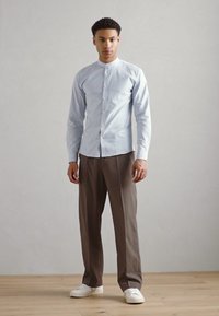 Camicia azzurro chiaro con colletto alla mandarino, abbinata a pantaloni marroni e sneakers bianche, in piedi su un pavimento di legno contro uno sfondo neutro.
