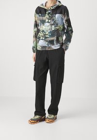 Giacca con cappuccio con stampa naturalistica in verde e blu, abbinata a pantaloni cargo neri. Le calzature presentano una combinazione di accenti verde oliva e arancione.