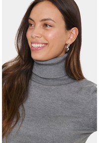 Pulover turtleneck cu model ribbed de culoare gri deschis, cu un design ajustat, având un guler înalt și o textură moale. Cercei argintii în formă de cerc sunt vizibili.