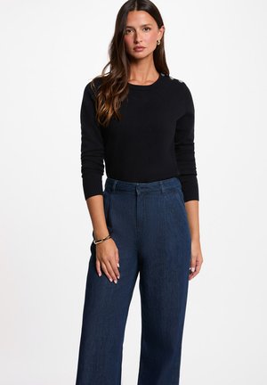 Mujer con cabello largo y castaño que lleva una camiseta negra de manga larga y unos vaqueros anchos de pierna con cintura alta y color azul oscuro, de pie sobre un fondo blanco.