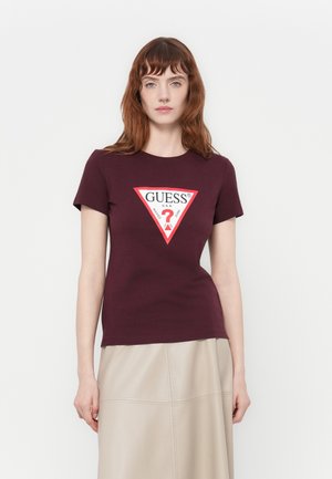 Donna che indossa una t-shirt attillata color bordeaux con un logo triangolare GUESS bianco e rosso, abbinata a una gonna beige su uno sfondo neutro.