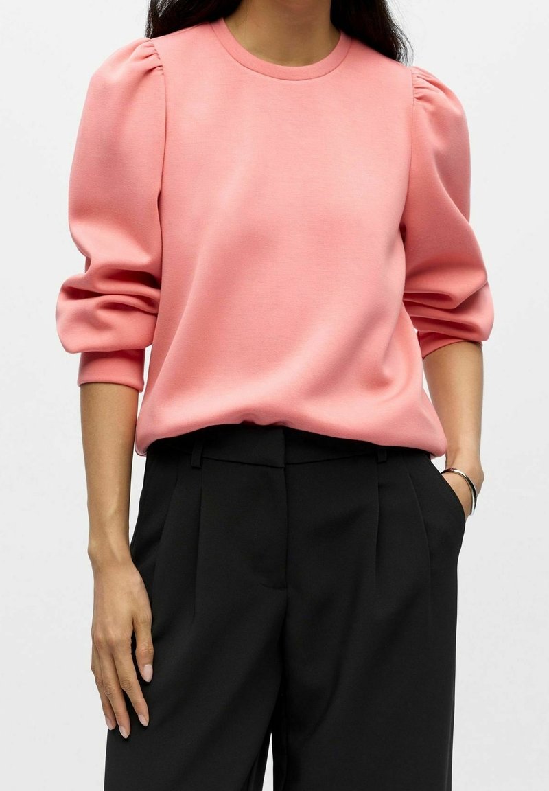 Femme portant un haut rose à manches bouffantes rentré dans un pantalon noir taille haute, une main dans la poche et un bracelet argenté au poignet.