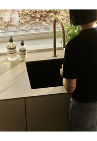 Lavello scuro integrato in un piano di lavoro beige con un rubinetto dorato. Due dispenser per sapone trasparenti e una pianta verde sono visibili nei dintorni.