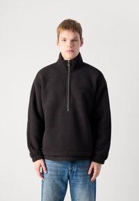 Pulover negru din fleece cu un guler înalt și închidere cu fermoar la un sfert. Prezintă o mică ecuson cu logo. Croială lejeră, textură moale și mâneci lungi.