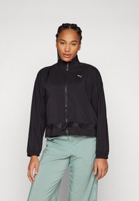 Chaqueta atlética negra con un alto cuello, cierre de cremallera en la parte delantera, dobladillo elástico y cordón ajustable. Presenta un pequeño logo en el pecho. Material ligero.