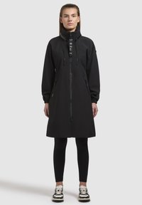 Cappotto nero di lunghezza al ginocchio con zip frontale, colletto alto e chiusura a toggles. Presenta polsini elasticizzati e tasche laterali con zip. Materiale leggero.