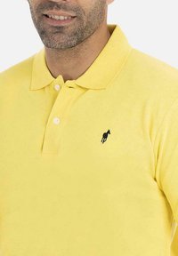 Polo shirt en coton jaune avec un col classique, portant un petit logo brodé noir sur la poitrine gauche. Texture douce et coupe standard.