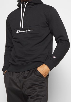 Zwarte hoodie met een ritskraag en een voorkantoor. Voorzien van het witte "Champion" logo op de borst en witte koordjes; geribde manchetten.