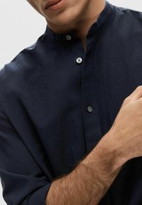 Chemise boutonnée bleu marine avec col montant, faite d'un tissu texturé. Présente des boutons en argent et des manches retroussées.