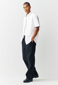 Chemise blanche à manches courtes boutonnée avec deux poches poitrine, associée à un pantalon noir ample et des chaussures foncées. Tissu lisse, coupe décontractée.