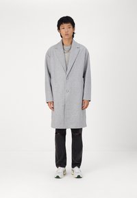 Cappotto di lana grigio con rever a vista, due bottoni e due tasche, indossato sopra un maglione a coste e abbinato a jeans neri strappati e sneakers.