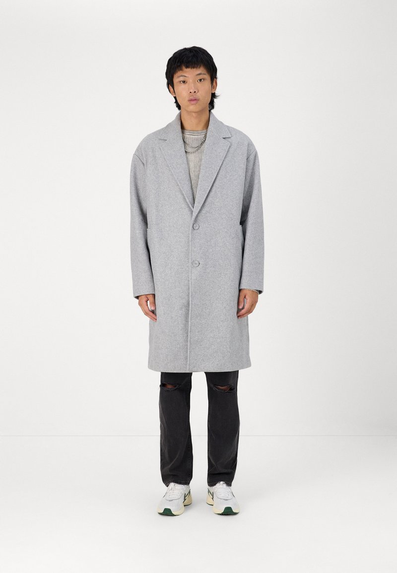 Cappotto di lana grigio con rever a vista, due bottoni e due tasche, indossato sopra un maglione a coste e abbinato a jeans neri strappati e sneakers.