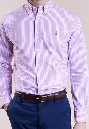 Mann trägt ein enganliegendes, lavendelfarbenes und weiß gestreiftes Hemd, das in blaue Hosen mit braunem Ledergürtel gesteckt ist.