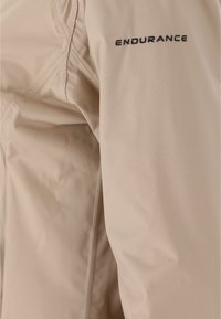 Beige jakke med en glat tekstur og synlige sømme. Logoet "ENDURANCE" er trykt med sort på ærmet.