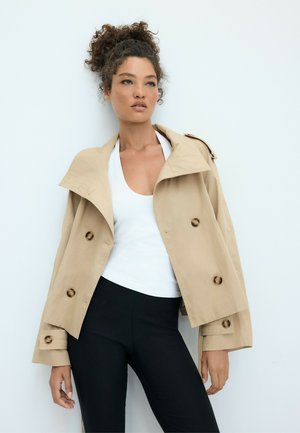 Femme aux cheveux bouclés portant un trench-coat beige sur un haut blanc et un pantalon noir, debout devant un fond clair uni.