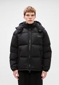 Crna puf jakna s patentnim zatvaračem sprijeda, podstavljenim dijelovima, bočnim zatvaračima i odvojivom kapuljačom. Ima logotip na prsima.