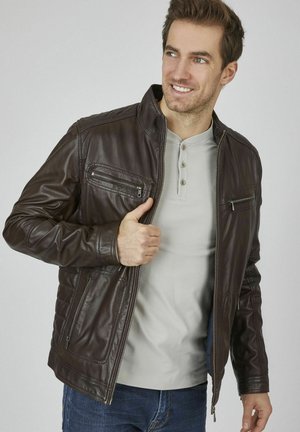 Chaqueta de cuero marrón con cierre frontal, cuello alto y varios bolsillos con cremallera. Lleva puesta una camiseta henley de color gris claro.