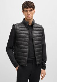 Black padded vest with a high collar, featuring a zip closure and horizontal quilted design. Worn over a black long-sleeve polo shirt.