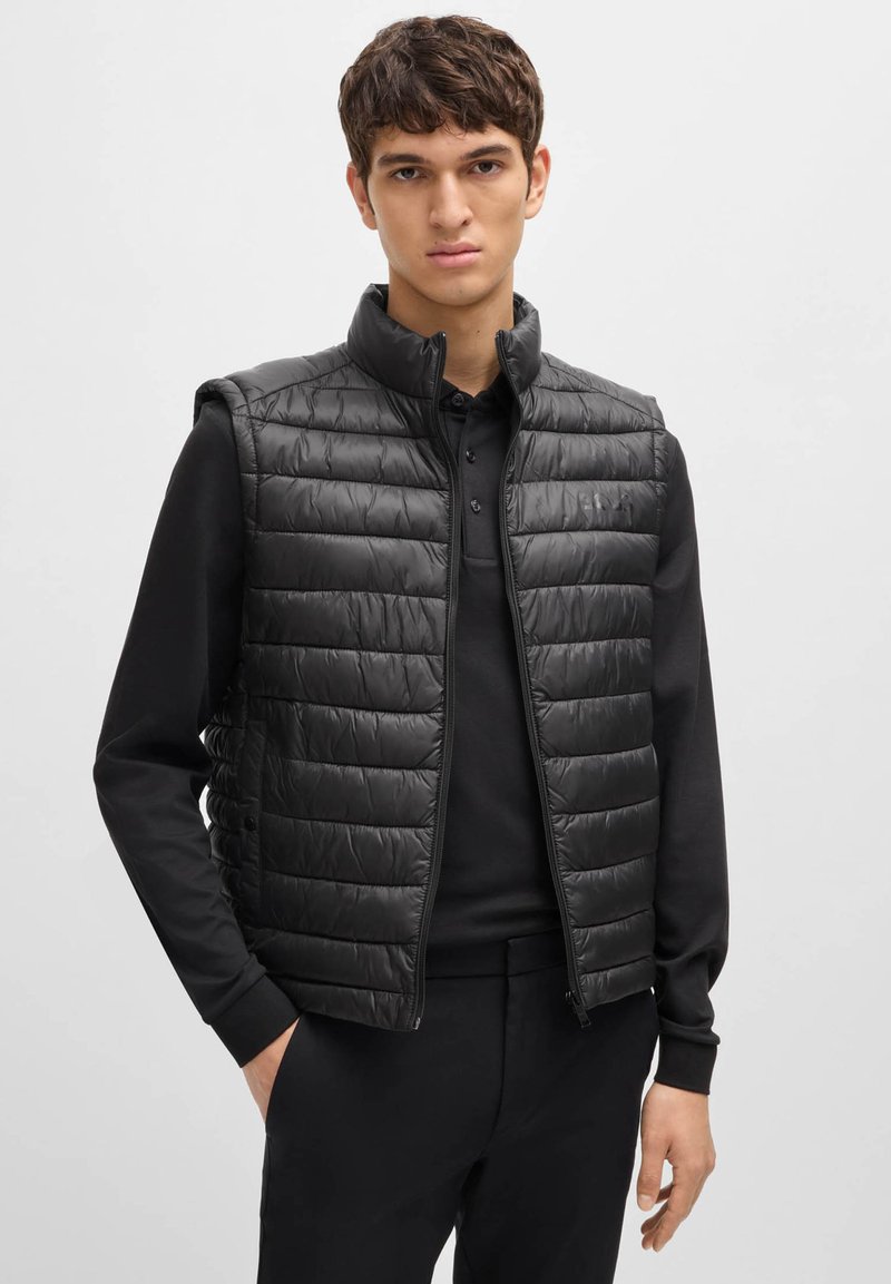 Black padded vest with a high collar, featuring a zip closure and horizontal quilted design. Worn over a black long-sleeve polo shirt.