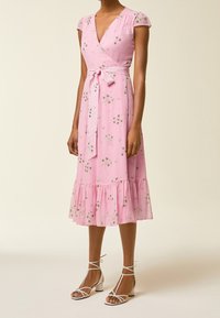 Rosa Blumen-Wickelkleid mit kurzen Ärmeln, einem Taillengürtel, einem rüschierten Saum und zarten Blumenmustern; kombiniert mit weißen Sandalen mit Knöchelriemen.