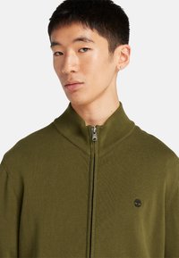 Olivgrön zip-up-tröja med hög krage och ribbat mönster. Har en liten svart logotyp på vänster bröst. Materialet verkar mjukt.