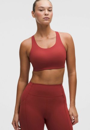 Frau trägt einen passenden roten Sport-BH und hochtaillierte Leggings, steht mit neutralem Gesichtsausdruck vor einem schlichten hellen Hintergrund.