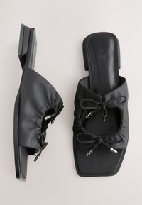 Schwarze Slip-On-Sandalen mit einer strukturierten Stoffoberfläche mit gerafften Details, quadratischer Zehenpartie und verstellbaren Bändern mit metallischen Akzenten.