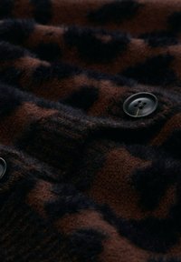Brauner gemusterter Cardigan mit schwarzen Flecken, gefertigt aus weichem Stoff. Verfügt über eine strukturierte Oberfläche und ein schwarzes Knopfdetail.