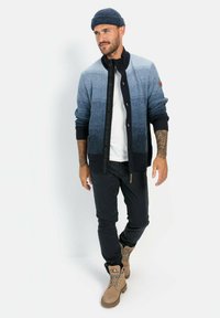 Strickjacke in blauem Farbverlauf mit Reißverschluss und gerippten Bündchen. Kombiniert mit einem weißen T-Shirt, dunklen Hosen und beigen Stiefeln.