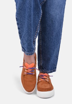 Persona che indossa jeans blu e scarpe da ginnastica arancione-marroni con suole bianche e lacci viola e arancioni, in piedi con i piedi vicini l’uno all’altro.