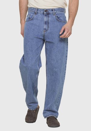 Uomo con jeans denim larghi azzurri chiari, scarpe marroni e una camicia bianca infilata, in piedi con una gamba leggermente avanti.