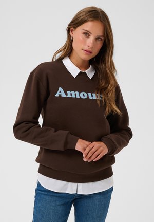 Jonge vrouw met lang bruin haar, die een bruine sweatshirt met de tekst "Amour", een wit overhemd met kraag en blauwe spijkerbroek draagt tegen een effen achtergrond.