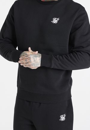 Personne portant un sweat-shirt noir et un pantalon avec des initiales blanches brodées sur la poitrine et la cuisse, mains jointes montrant des tatouages.