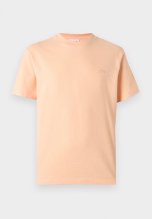 Jednoduché svetlo broskyňové tričko s krátkym rukávom a okrúhlym výstrihom, s malým vyšívaným logom na ľavej strane hrudníka, zobrazené na bielom pozadí.