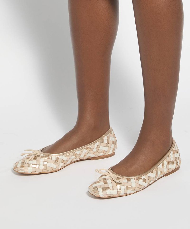 Jambes brunes portant des ballerines tissées beige et blanche avec de petits nœuds sur fond blanc.