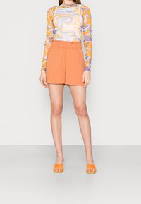 Langärmelig top in meerdere kleuren met draaipatronen, gecombineerd met oranje korte broek met elastische tailleband en oranje instapsandalen. Gladde textuur.