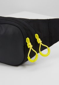 Bolsa de cintura de tela negra con dos tiradores de cremallera amarillos que cuentan con empuñaduras de cordón en una superficie clara.