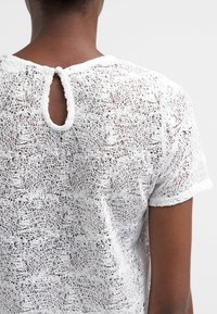 Top corto bianco a maniche corte con un motivo di pizzo testurizzato, dettaglio a forma di chiave circolare sul retro e tessuto morbido e leggero.