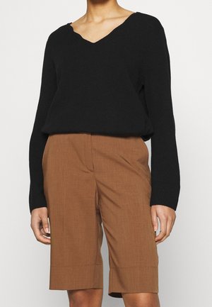 Osoba ubrana w czarny sweter z długim rękawem i dekoltem w serek oraz brązowe, wysokie szorty sięgające kolan, stojąca na tle jednolitego tła.