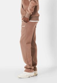 Pantalones de chándal de algodón marrón con bolsillos laterales y un logotipo blanco en la pierna izquierda. Combinados con zapatillas deportivas blancas. Textura suave y ajuste holgado.