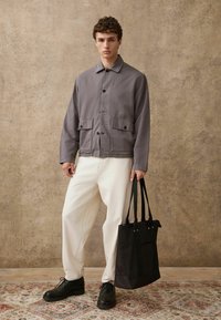 Szara, lekka kurtka z guzikami z przodu i dwiema kieszeniami; zestawiona z białymi, luźnymi spodniami i czarnymi sznurowanymi butami; trzyma czarną torbę.