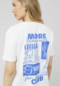 T-shirt en coton blanc avec un imprime graphique bleu représentant une scène de café, des tasses de café et le texte "Plus de plaisir pour le Brunch Club." Manches courtes.